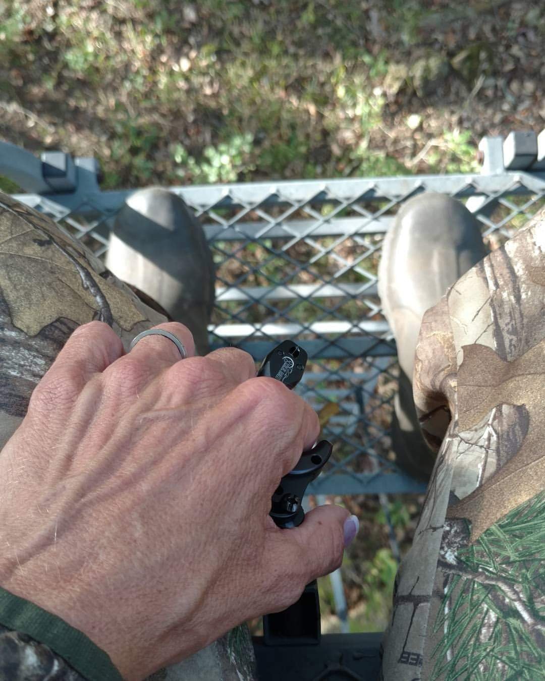 woman sitting in her treestand looking down at how high up she is with a release in her hand